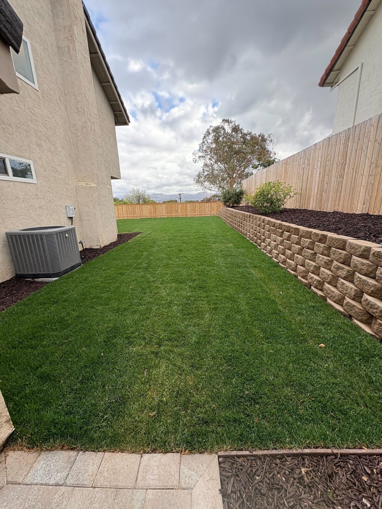 Completed narrow backyard landscape showing a full artificial turf lawn with a segmental retaining wall and a small interlocking paver patio at the foreground. Mulch beds border the wall and house, with a wood privacy fence and HVAC unit visible — overall appears as a finished installation.