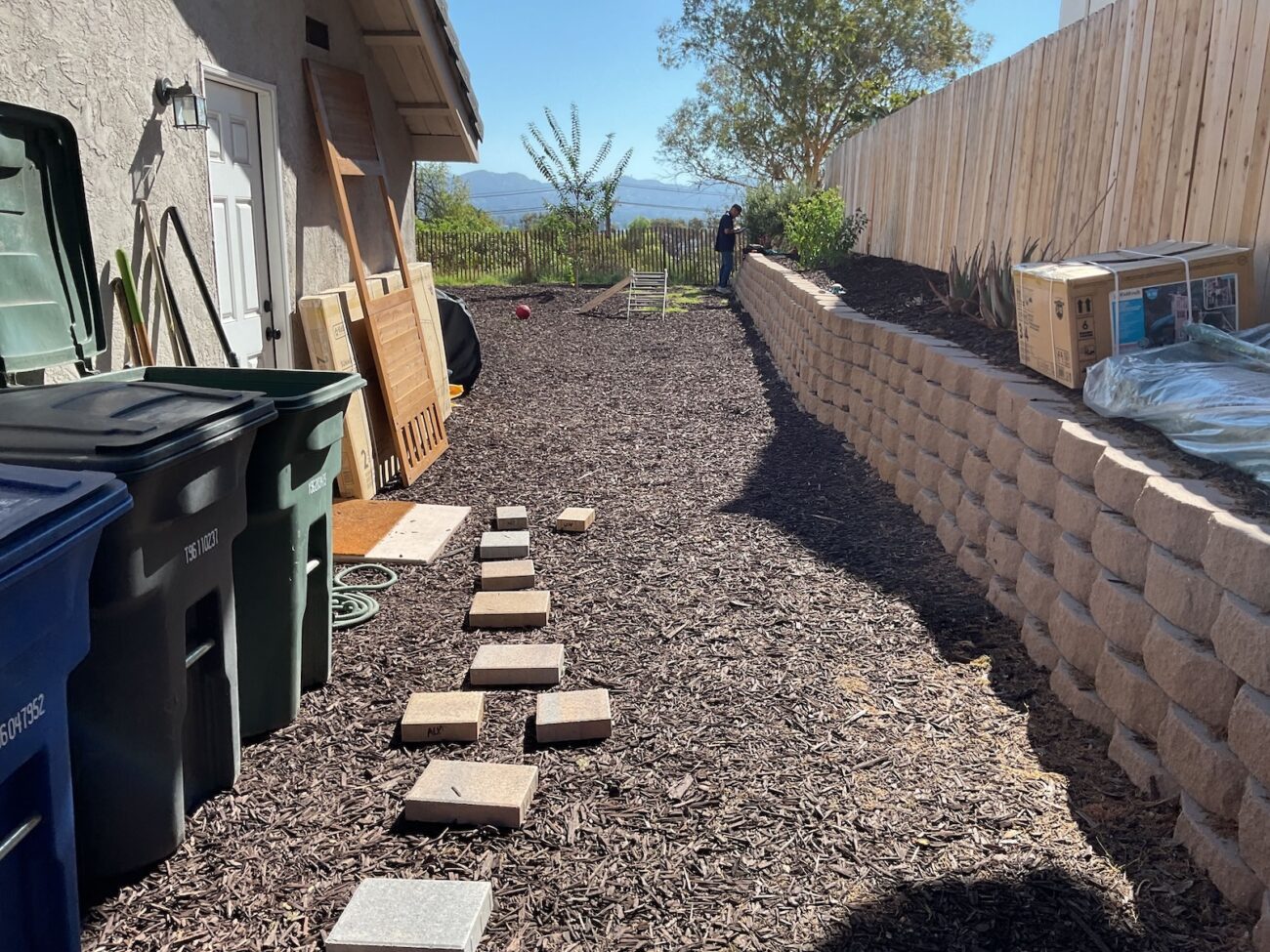 Side yard of a residential property showing a segmental concrete retaining wall with a mulch-covered area and a temporary line of stepping stone pavers leading toward the far end. Construction materials and trash bins are staged along the house and a worker is visible near the top of the wall, indicating ongoing installation.