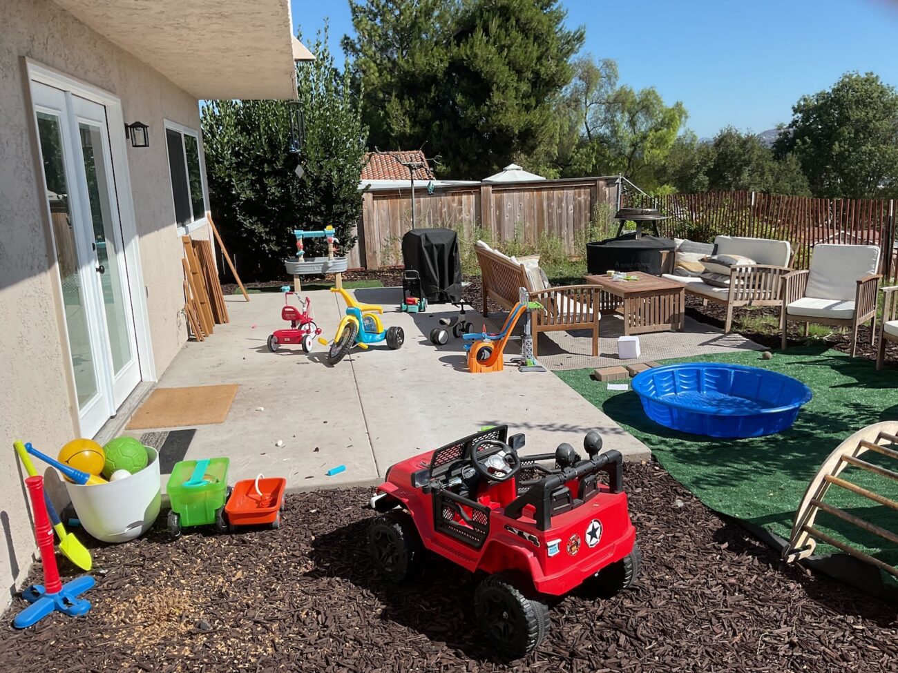 Concrete patio slab with outdoor seating and living area adjacent to a recently installed artificial turf section and mulched planting bed. Children's toys, a plastic kiddie pool and a ride-on toy on the turf indicate the backyard landscape is in use post-installation.