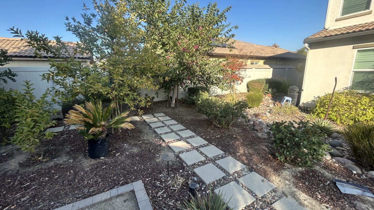 Narrow side yard alongside a residence with mulch installed over planting bed areas and a soil strip remaining along the access path. A run of concrete stepping stones leads toward the rear, a gas meter is mounted on the house exterior, and refuse bins are staged at the far end. Image indicates landscape prep in progress prior to final surfacing.