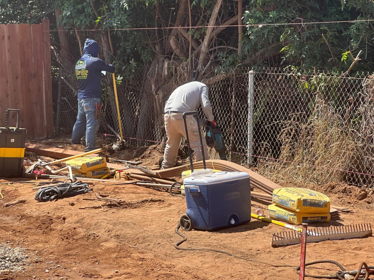 Two workers installing drain pipe and setting fence posts on a red-dirt job site with Quikrete bags and a cooler nearby.