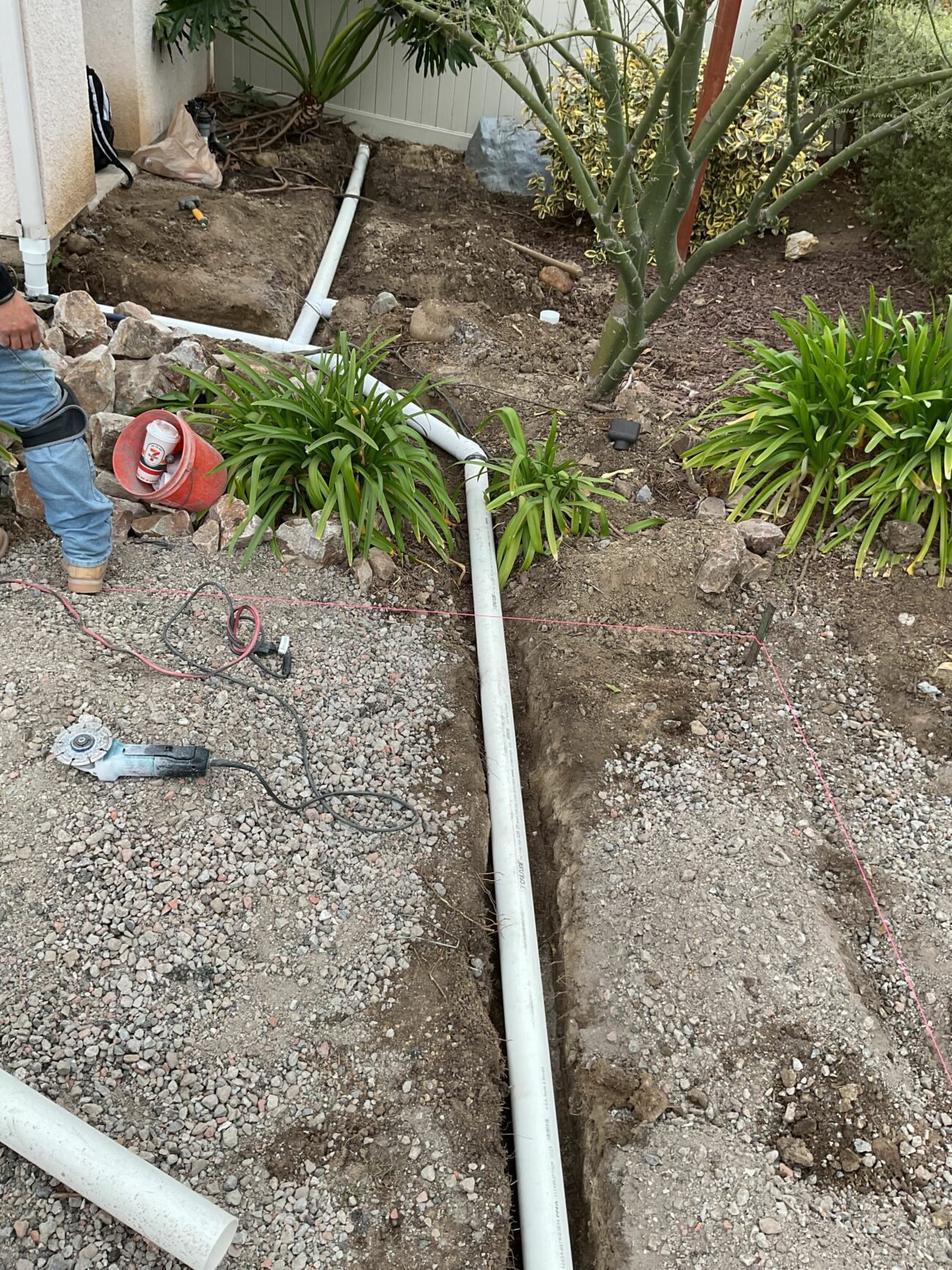 Worker laying white PVC drainage pipe in a shallow trench through a residential front yard garden bed.