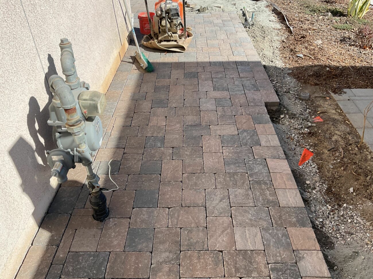 Newly laid dark brown tumbled cobble pavers along a stucco wall with a gas meter, construction equipment visible in backgr...
