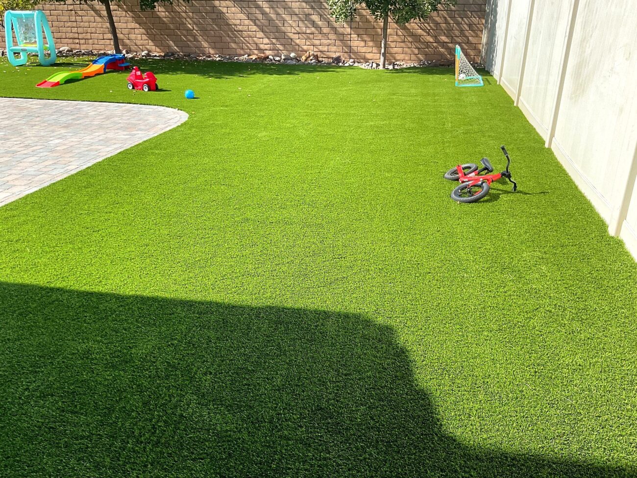 Residential backyard showing newly installed artificial turf abutting an interlocking paver patio. Clean turf field with visible edge detail and a privacy wall and planting bed along the perimeter; children's toys and a small bike on the turf indicate final site use.