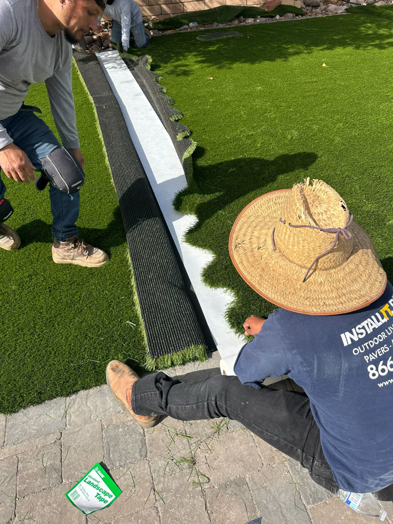 Two workers joining seams of synthetic turf rolls using landscape tape and a seaming strip on a residential lawn.