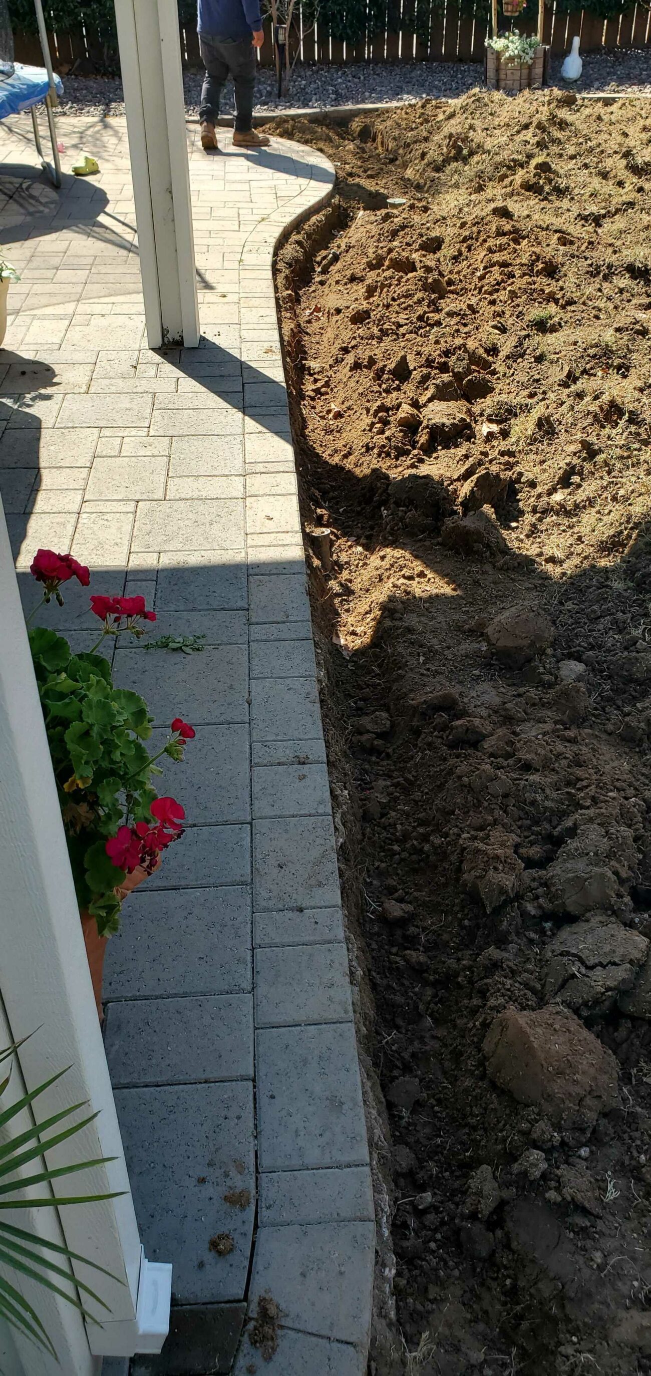 Excavated backyard area next to a paver patio edge, with loose dirt and a shallow trench running along the border.