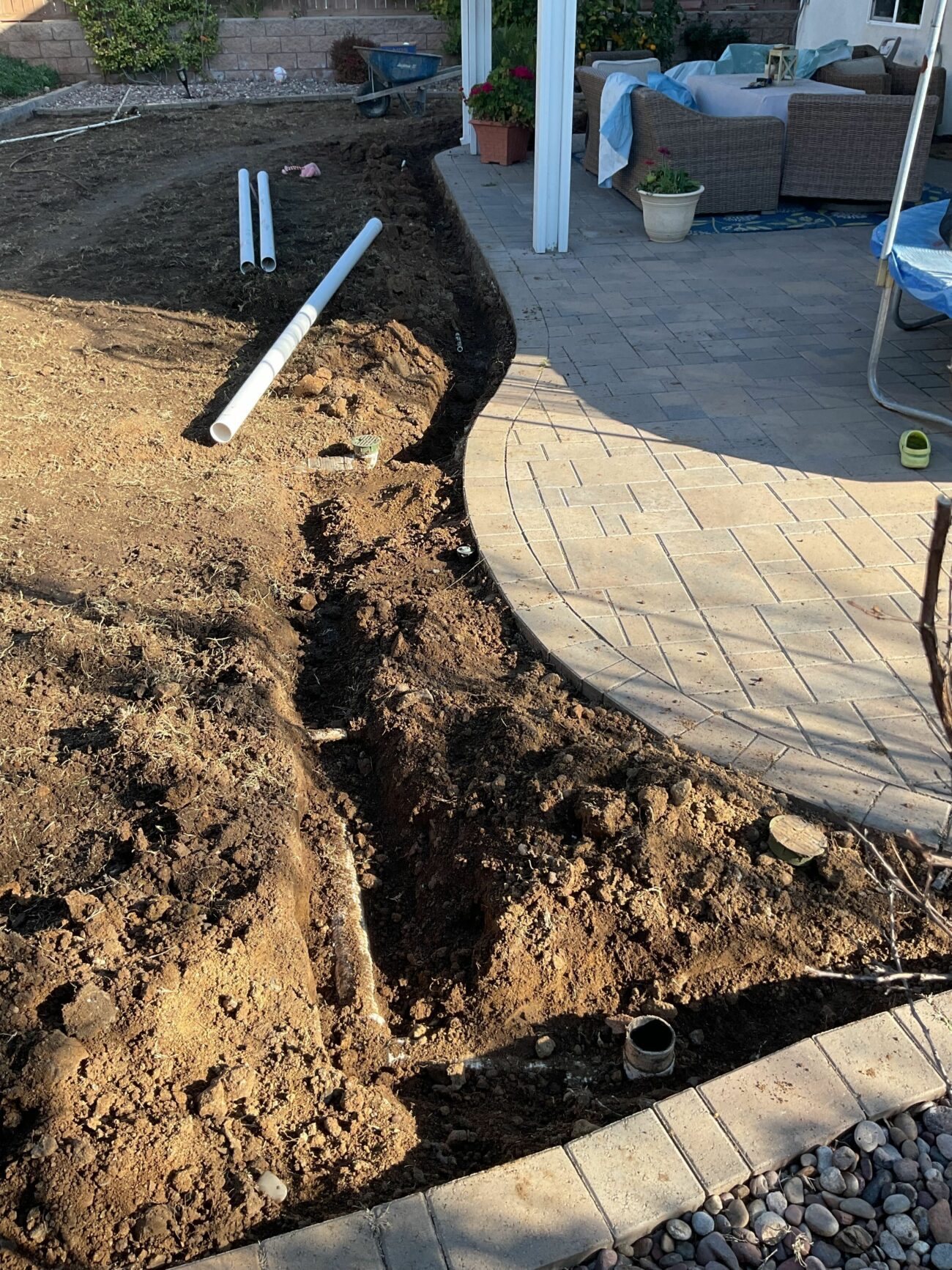 Trench dug along curved paver patio edge with white PVC drain pipes laid nearby, ready for installation.