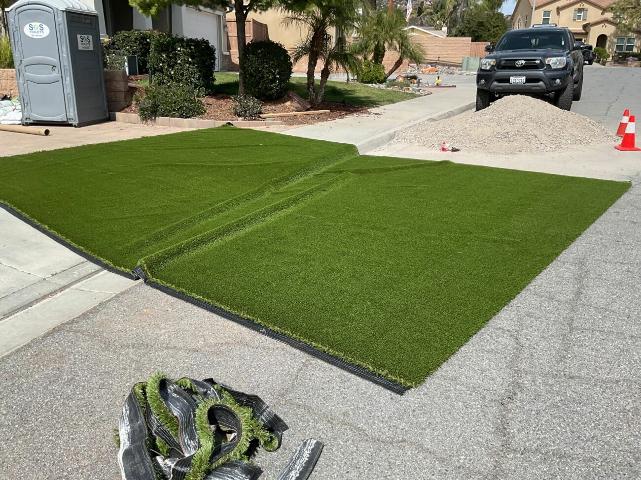 Two sections of artificial turf laid flat on a residential driveway in Lakeside, with a gravel pile and portable toilet ne...