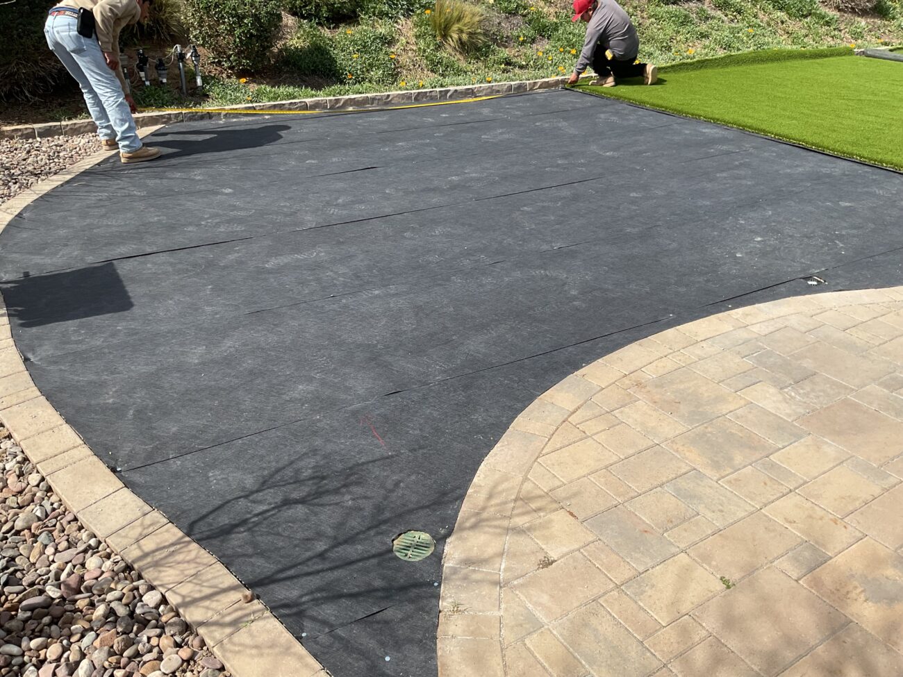 Two workers unrolling artificial turf over a dark weed barrier on a curved backyard patio bordered by pavers and river rock.