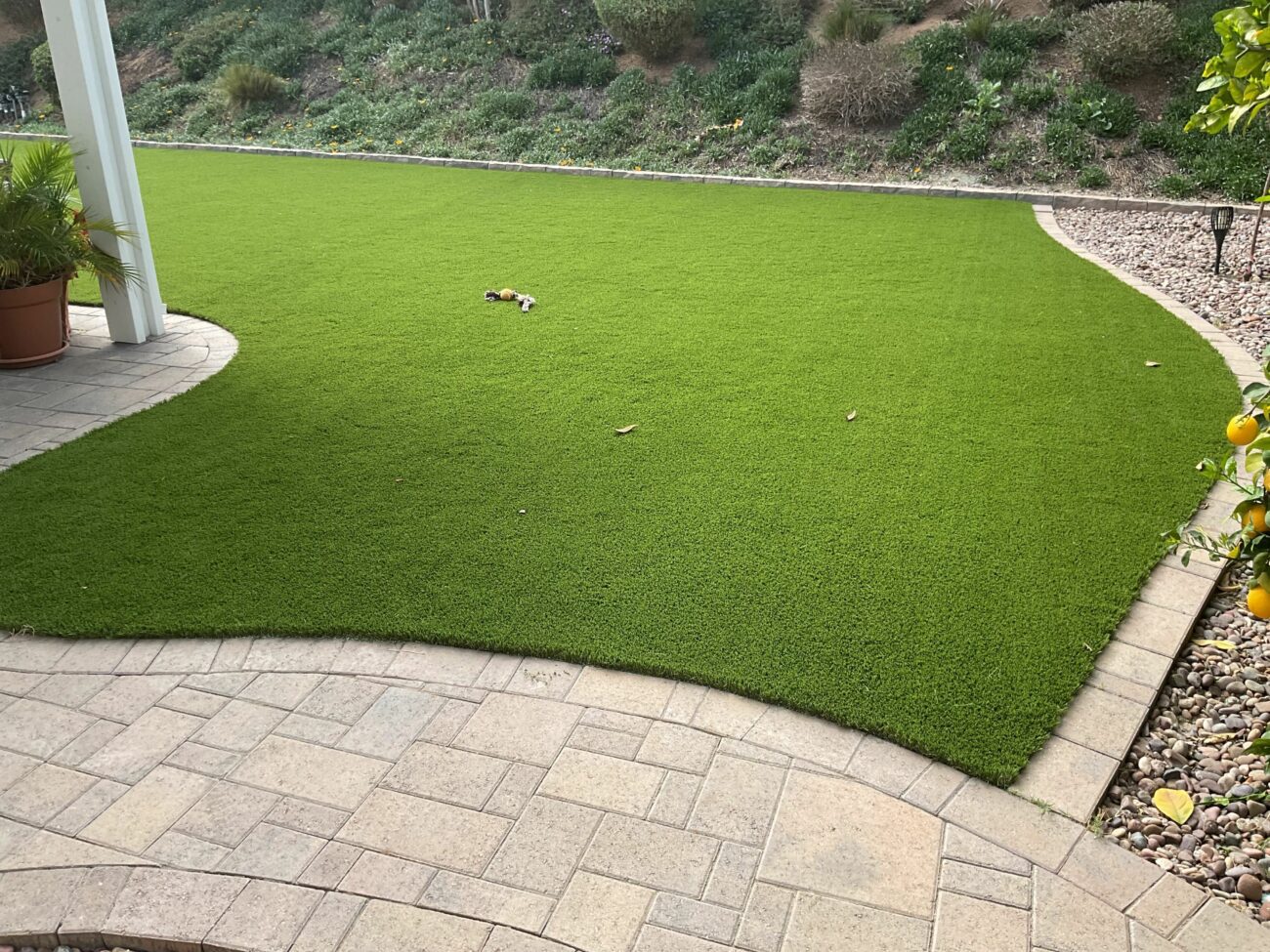 Lush artificial turf lawn bordered by curved stone pavers, a gravel bed, and a retaining wall with a hillside beyond.