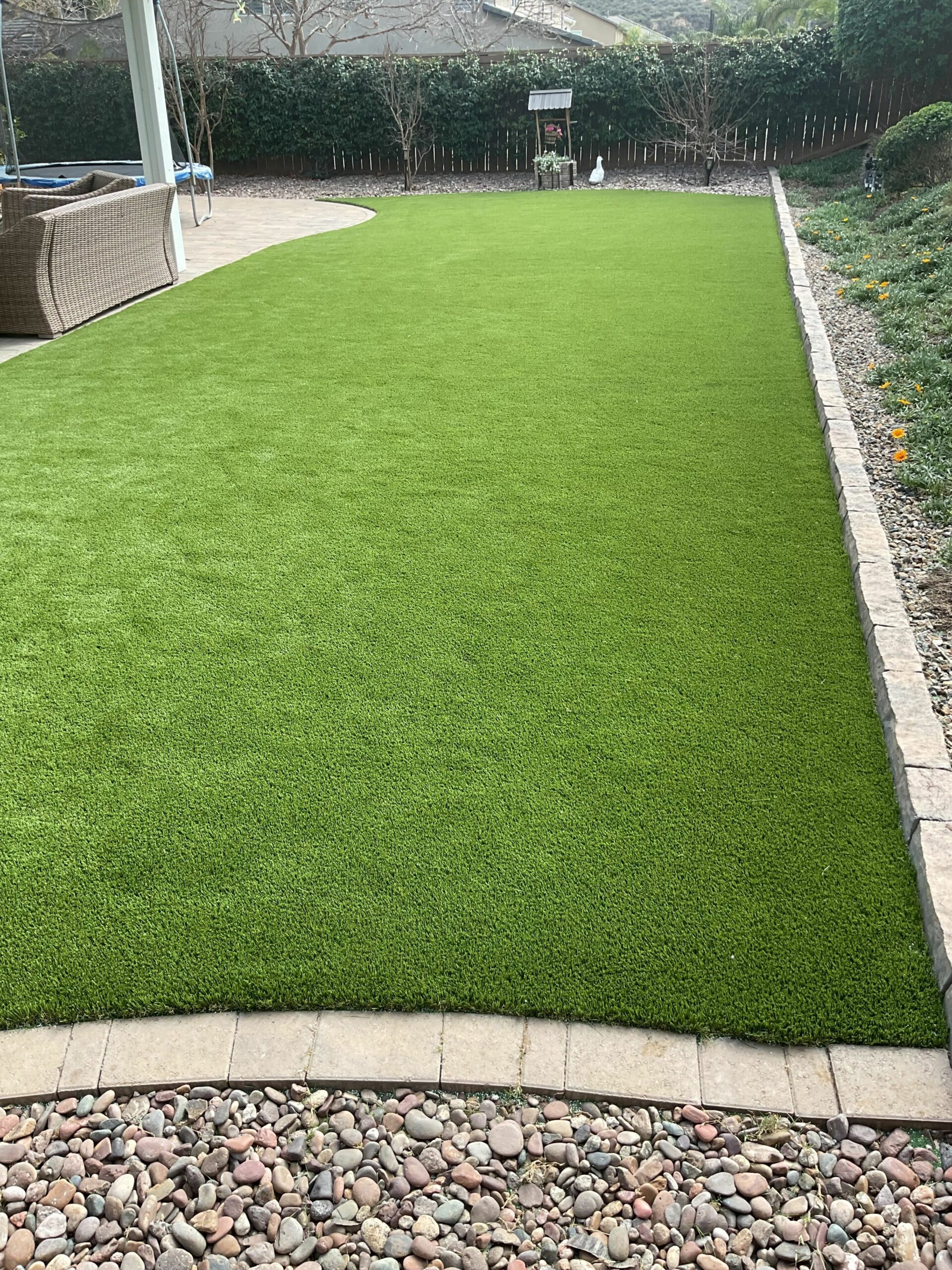 Lush artificial turf lawn bordered by stone pavers and river rock in a Lakeside backyard, with patio seating and wood fenc...