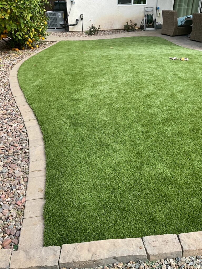 Curved artificial turf lawn edged with concrete pavers and river rock, with a citrus tree and AC unit visible in the backy...