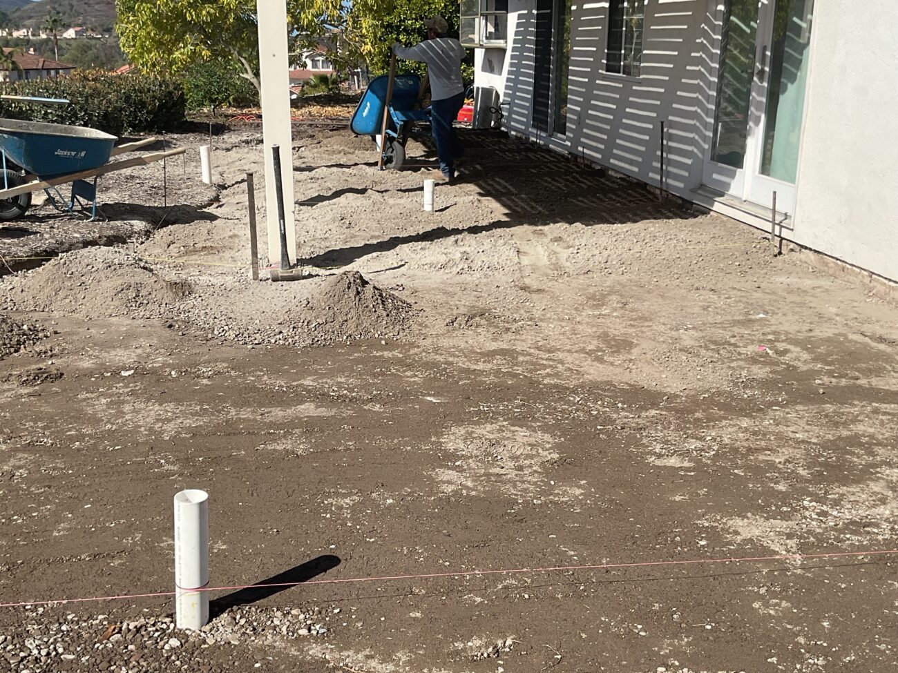 Graded dirt backyard beside a modern home with PVC stub-outs, string lines, and a wheelbarrow during site prep.