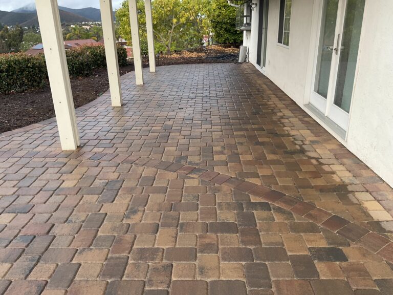 Herringbone-pattern brick pavers covering a covered patio alongside a white stucco home with mountain views beyond.