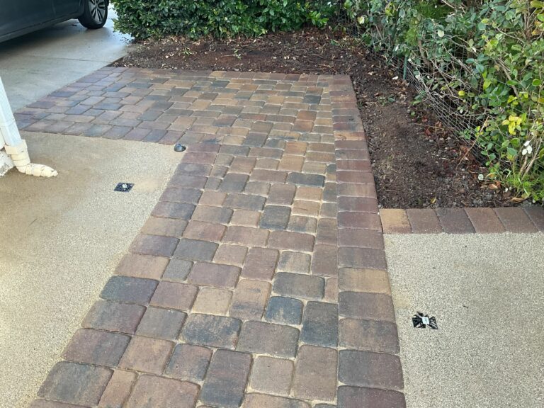 Brick-tone clay pavers forming an L-shaped walkway set into an exposed-aggregate concrete driveway, flanked by shrub beds.