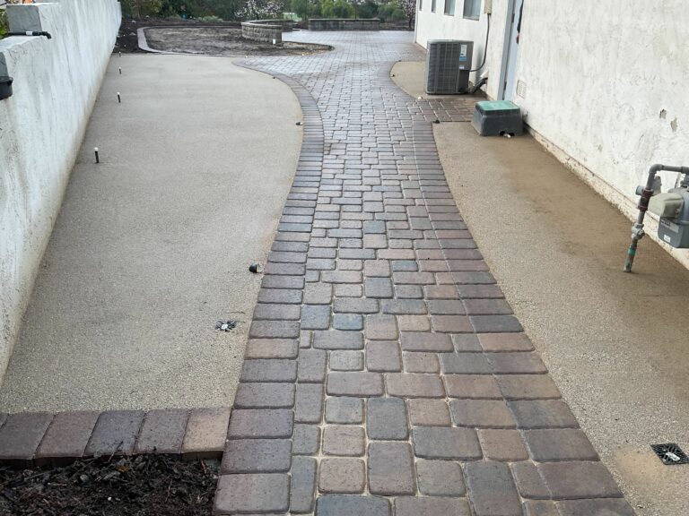 Curved cobblestone paver path flanked by stabilized decomposed granite on both sides, running alongside a white stucco bui...