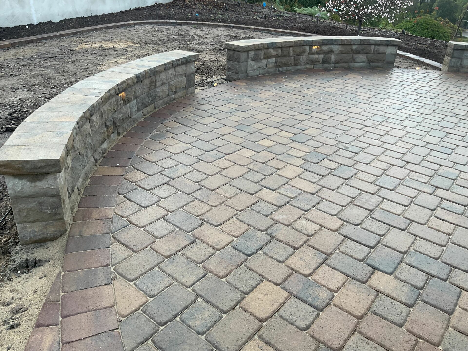 Curved segmental seat wall with cap stones and small path lights bordering a herringbone-pattern concrete paver patio.