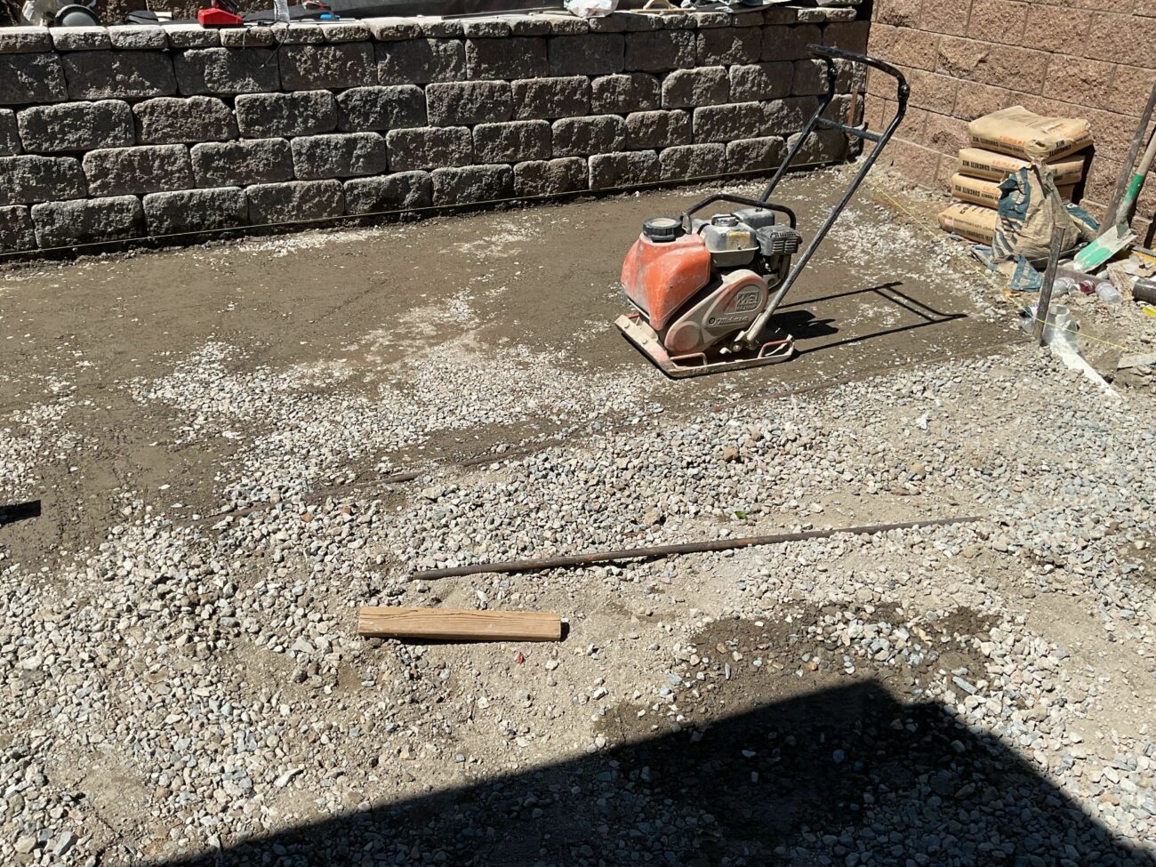 Plate compactor sitting on compacted gravel base in a backyard construction area with a block retaining wall behind it.
