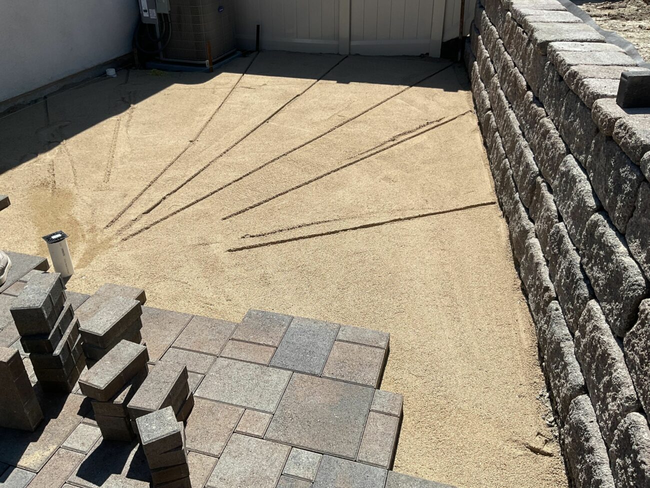 Compacted gravel base with radial string layout lines marked across it, bordered by a segmental retaining wall and stacked...