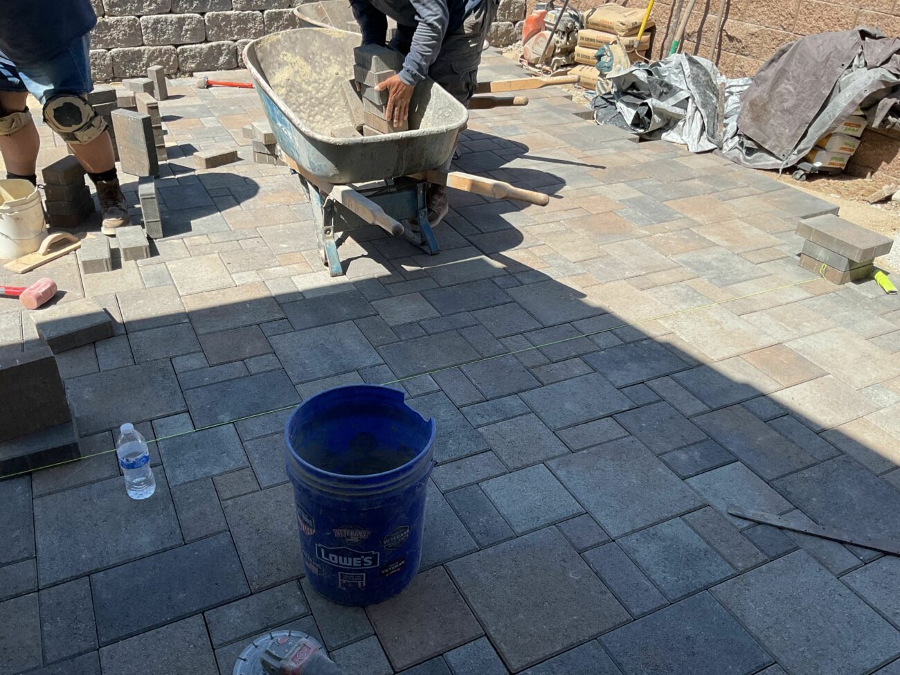 Workers laying interlocking pavers in a modular pattern, with a loaded wheelbarrow and stacked paver materials nearby.