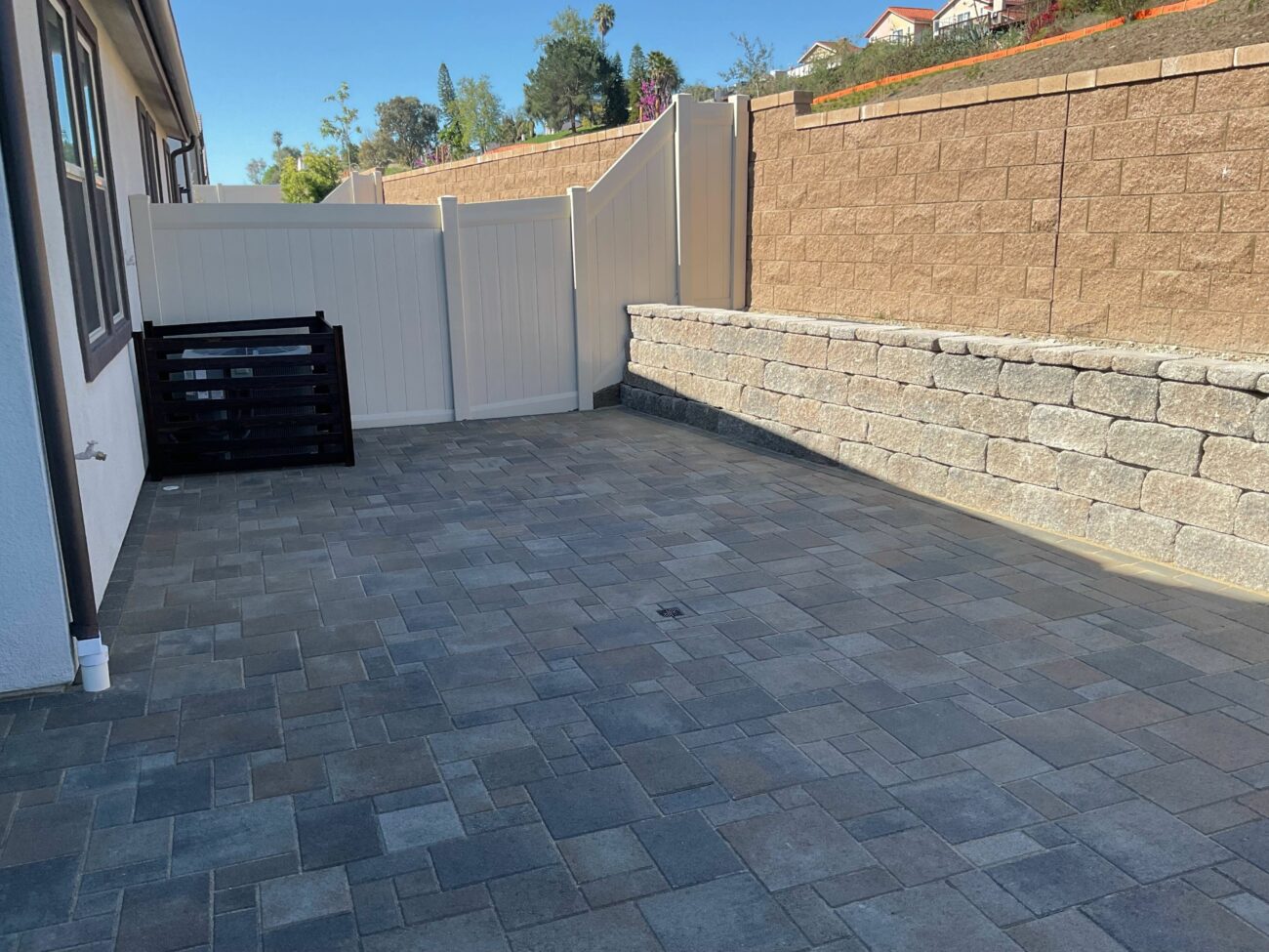 Finished installation of multi-size concrete pavers forming a residential patio area adjacent to the house. A segmental retaining wall and a white vinyl fence are installed along the perimeter and a surface drain is visible in the paved area, indicating final site grading and drainage integration.
