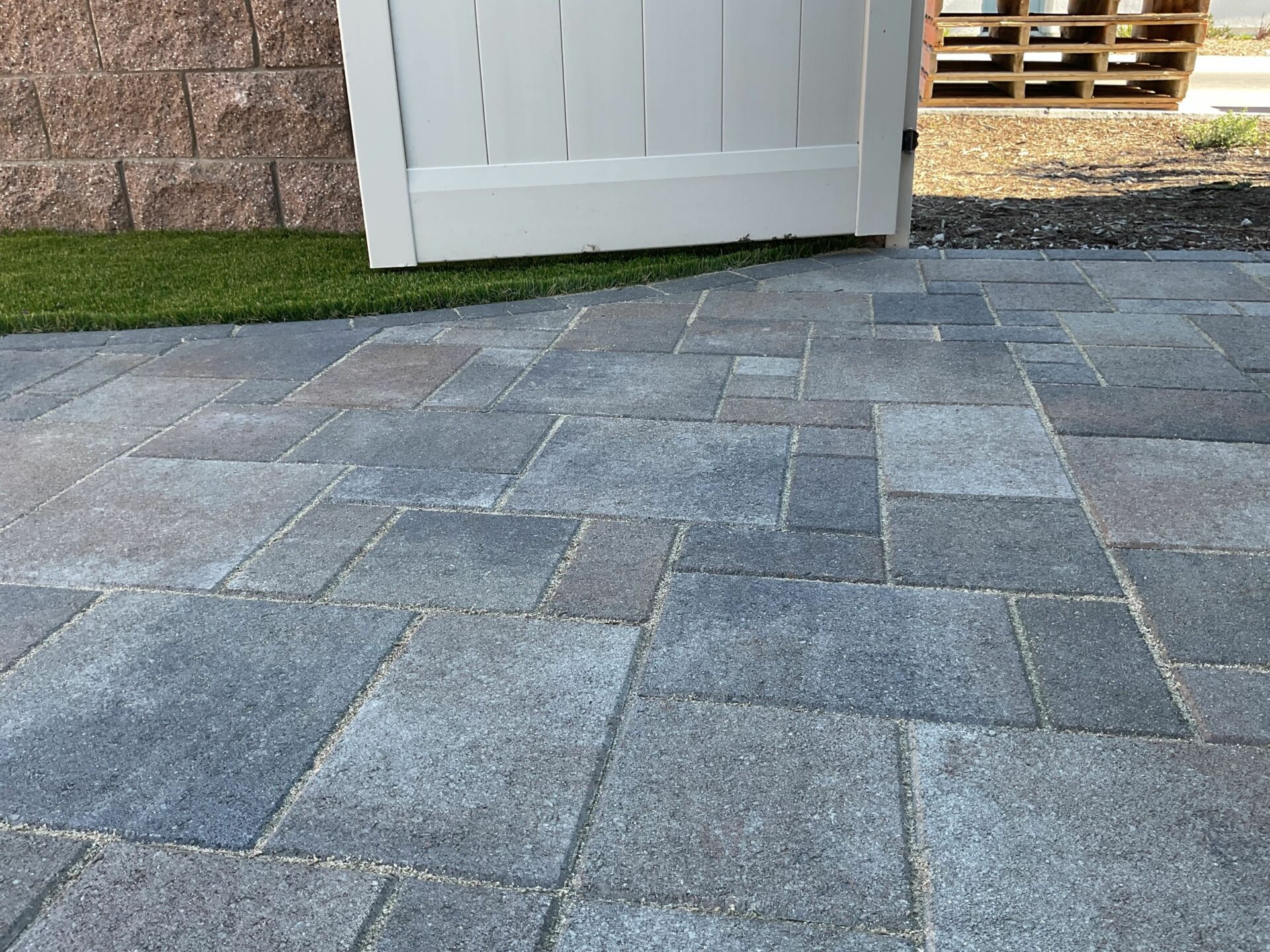 Close-up of a gray interlocking concrete paver patio in a modular ashlar pattern beside a white gate and green turf.