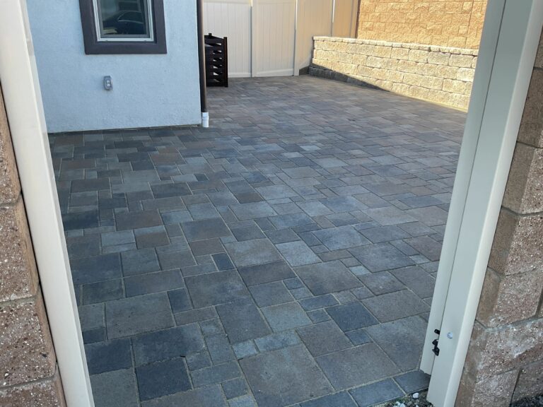 Gray interlocking pavers in a modular ashlar pattern fill a side-yard patio viewed through an open door.