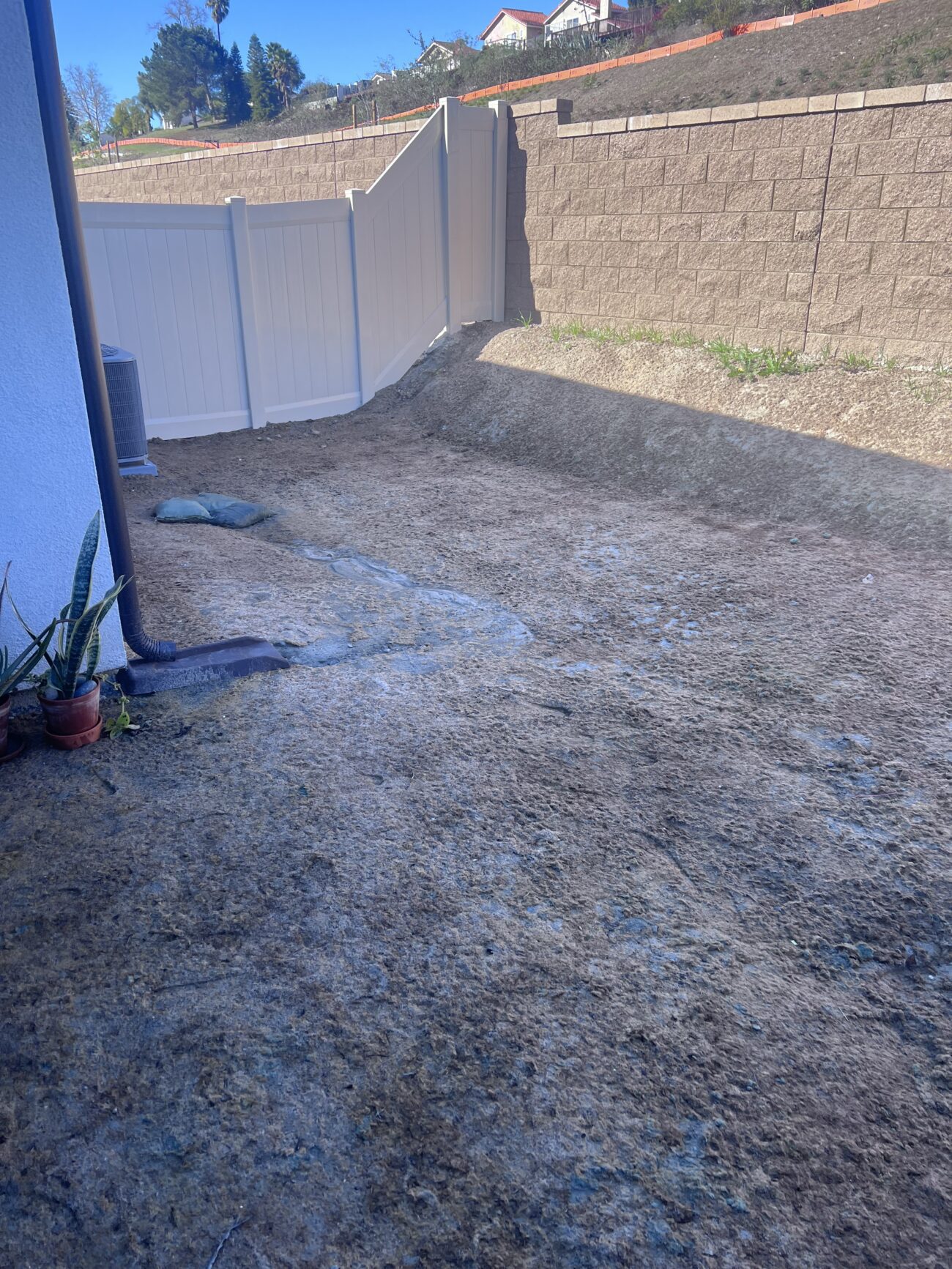 Sloped residential backyard with a segmental concrete retaining wall and vinyl fence; the yard subgrade is bare soil covered with erosion-control straw/mulch and looks prepared for future landscape or hardscape work. Small potted plants sit against the house exterior at the left, and there are no pavers or turf installed yet.