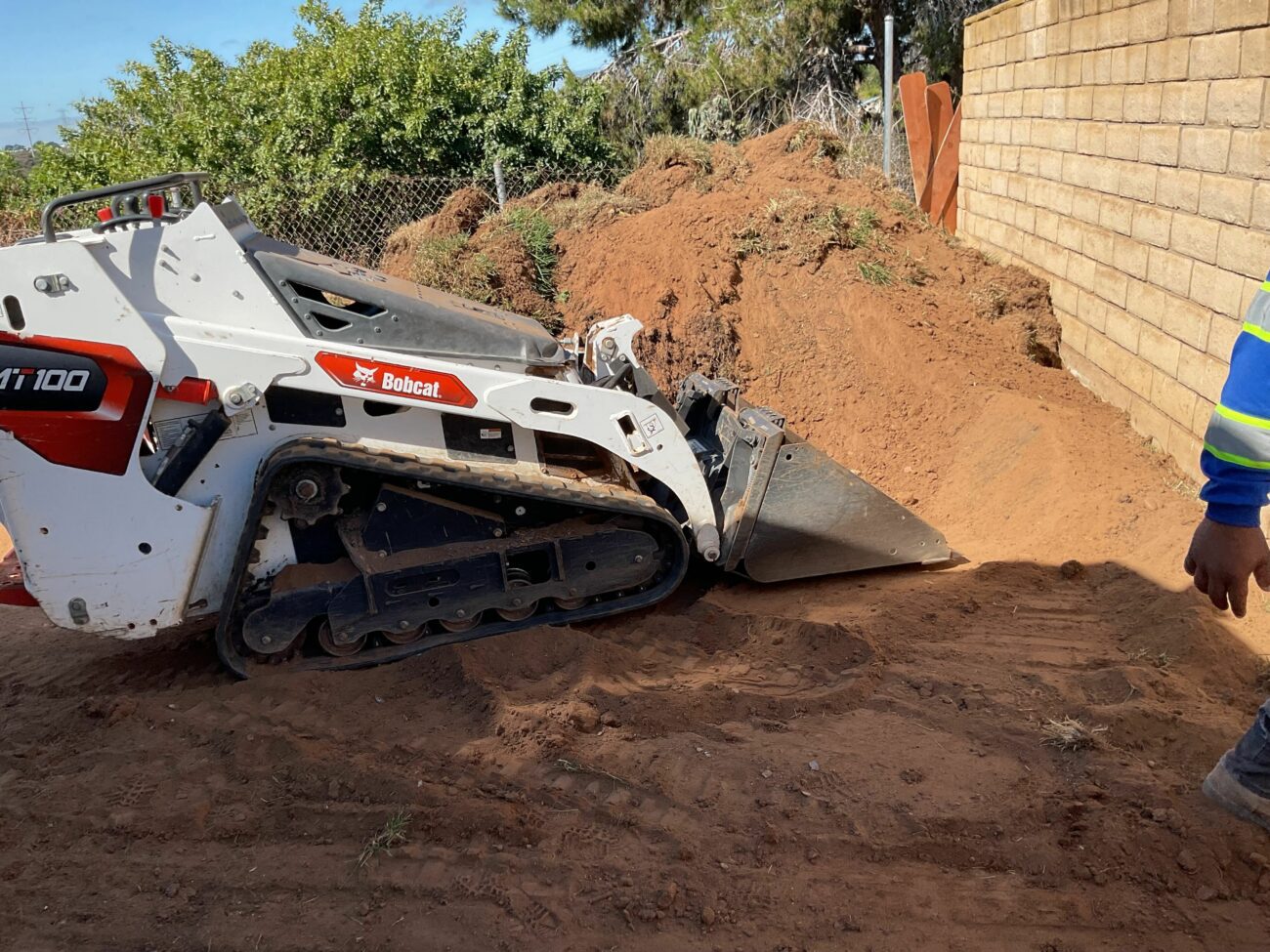Bobcat MT100 compact track loader digging into a dirt mound along a block wall during site grading.