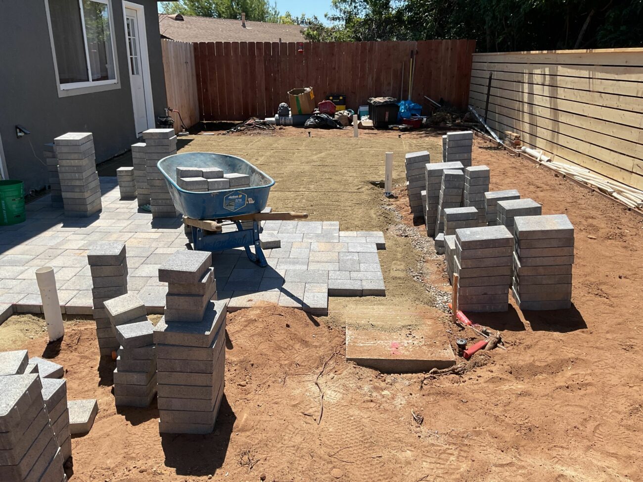 Backyard patio under construction with stacked concrete pavers, a blue wheelbarrow, and exposed red dirt in San Diego.