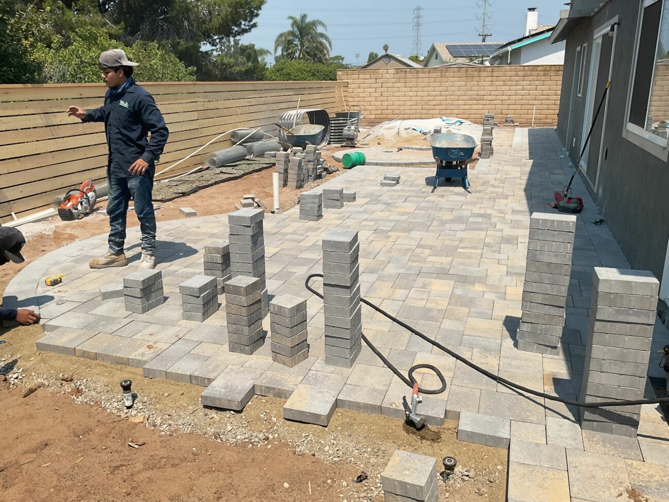 Worker stands on a partially laid gray concrete paver patio in a backyard, with stacked pavers and tools scattered around.