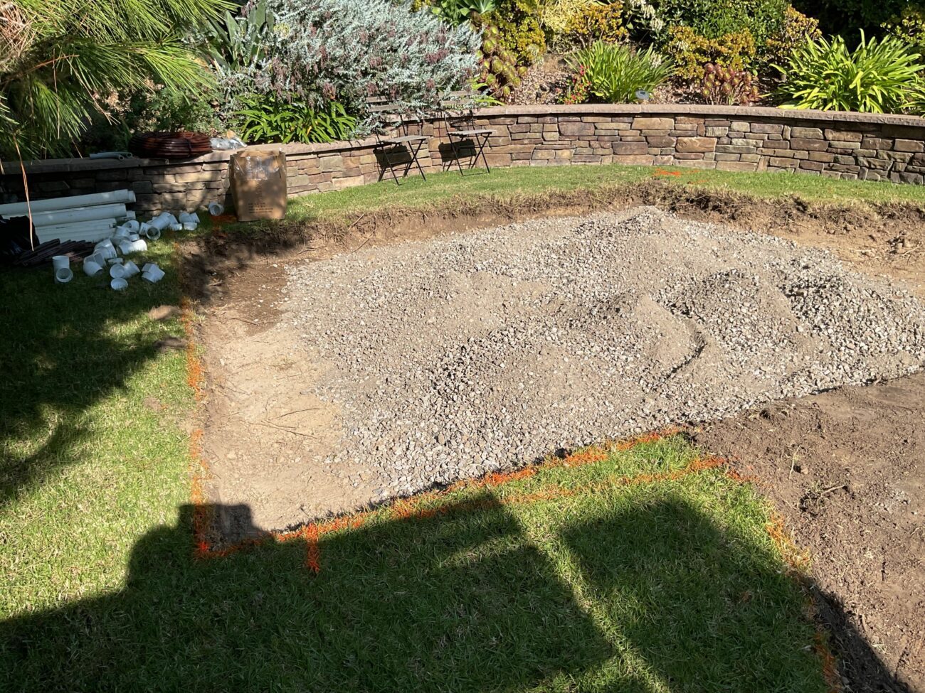 Excavated backyard area with crushed gravel base layer spread across the dig, orange spray paint marking patio boundaries...