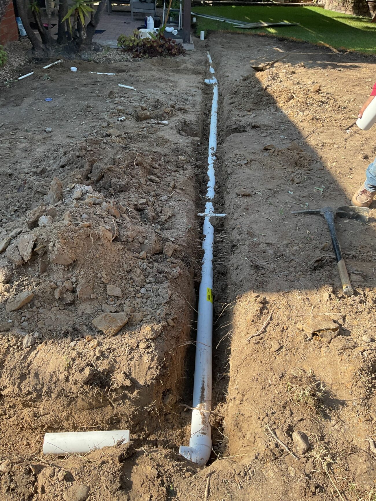 PVC drain pipe laid in a narrow trench across a dirt yard, with a pickaxe resting on the trench edge.