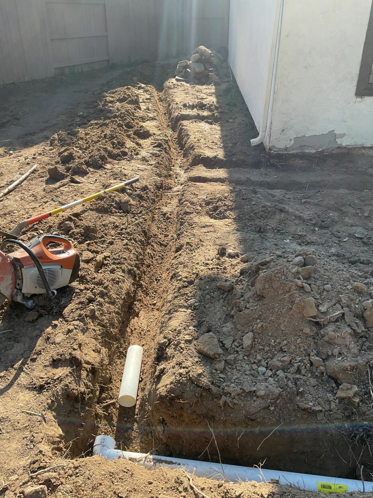 Narrow trench dug along a house wall with white PVC drain pipe laid in it and a trenching machine resting nearby.