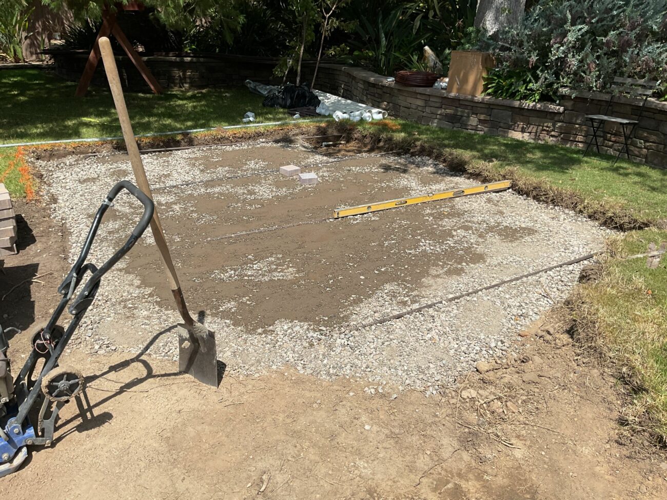 Gravel base compacted and leveled in a backyard excavation, with a spirit level, shovel, and hand truck visible nearby.
