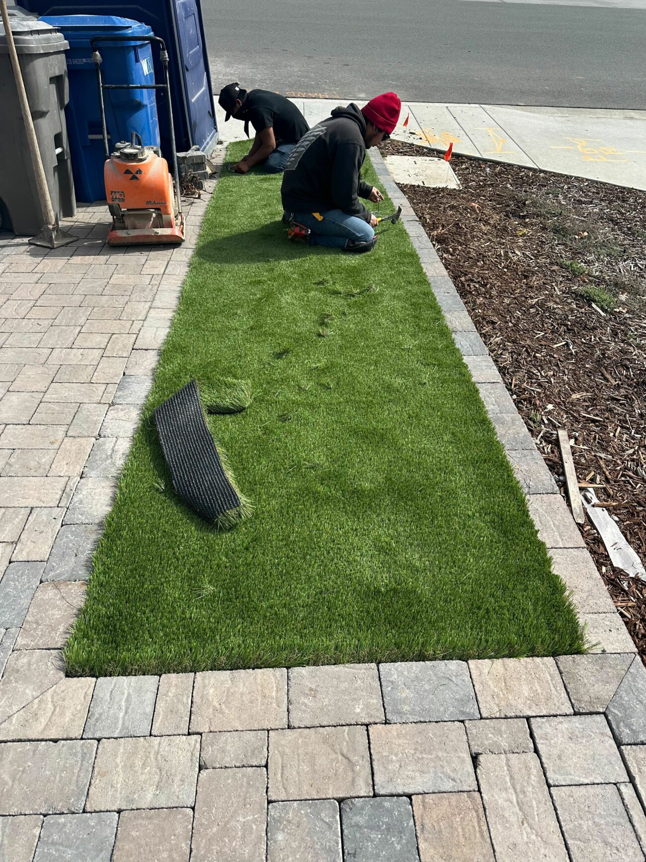 Two workers kneeling to install a strip of synthetic turf between stone pavers and a mulched planting bed.