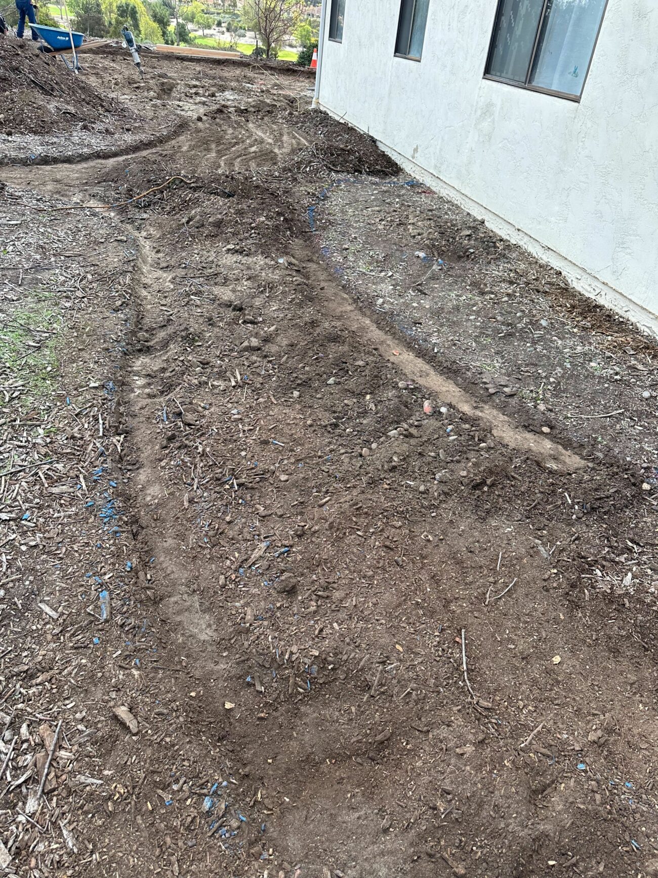 Freshly trenched drain line channel running alongside a stucco home's foundation through bare, disturbed soil.