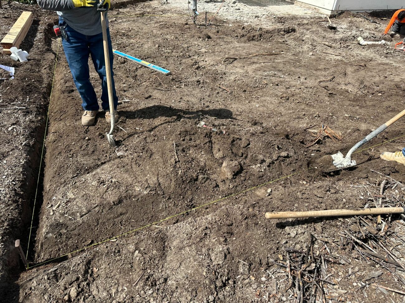 Worker using a fork to grade and level a dirt construction site, with layout strings marking the perimeter.