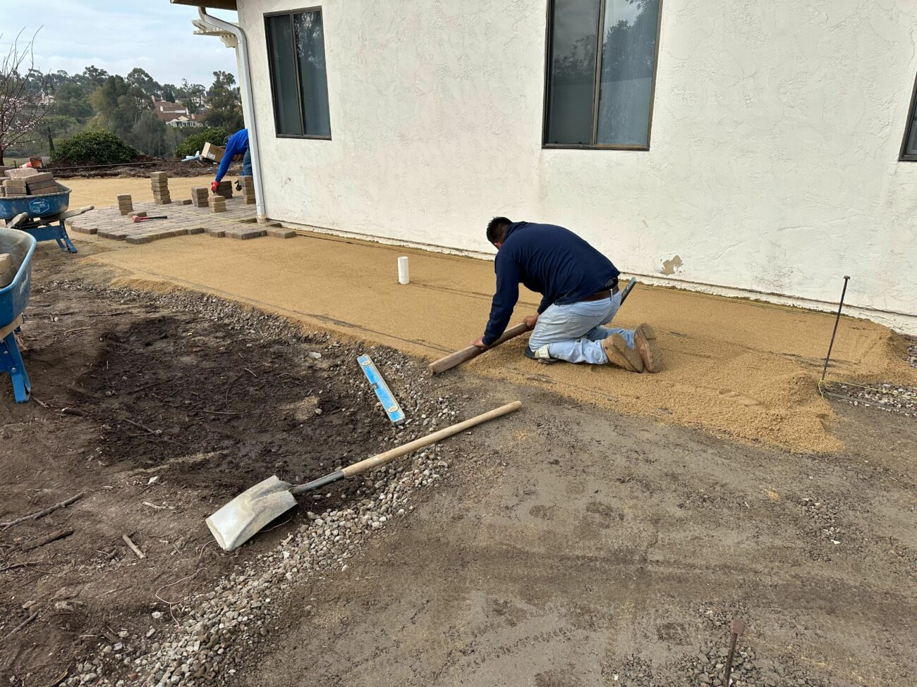Worker on knees screeding a sand base alongside a house foundation while a second worker lays pavers in the background.