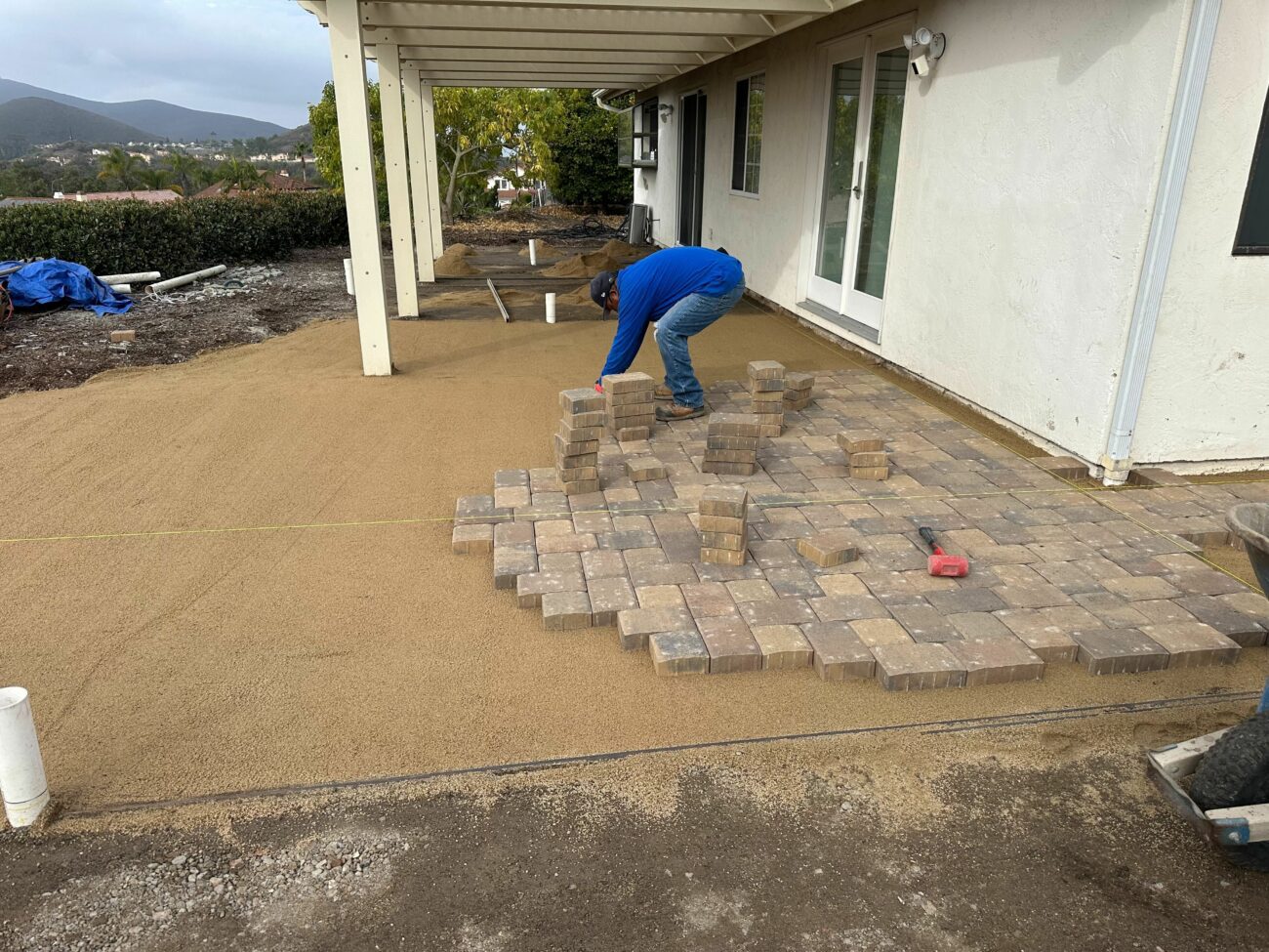 Worker placing ORCO Holland pavers on a sand base beside a stucco house, with mountains visible in the background.