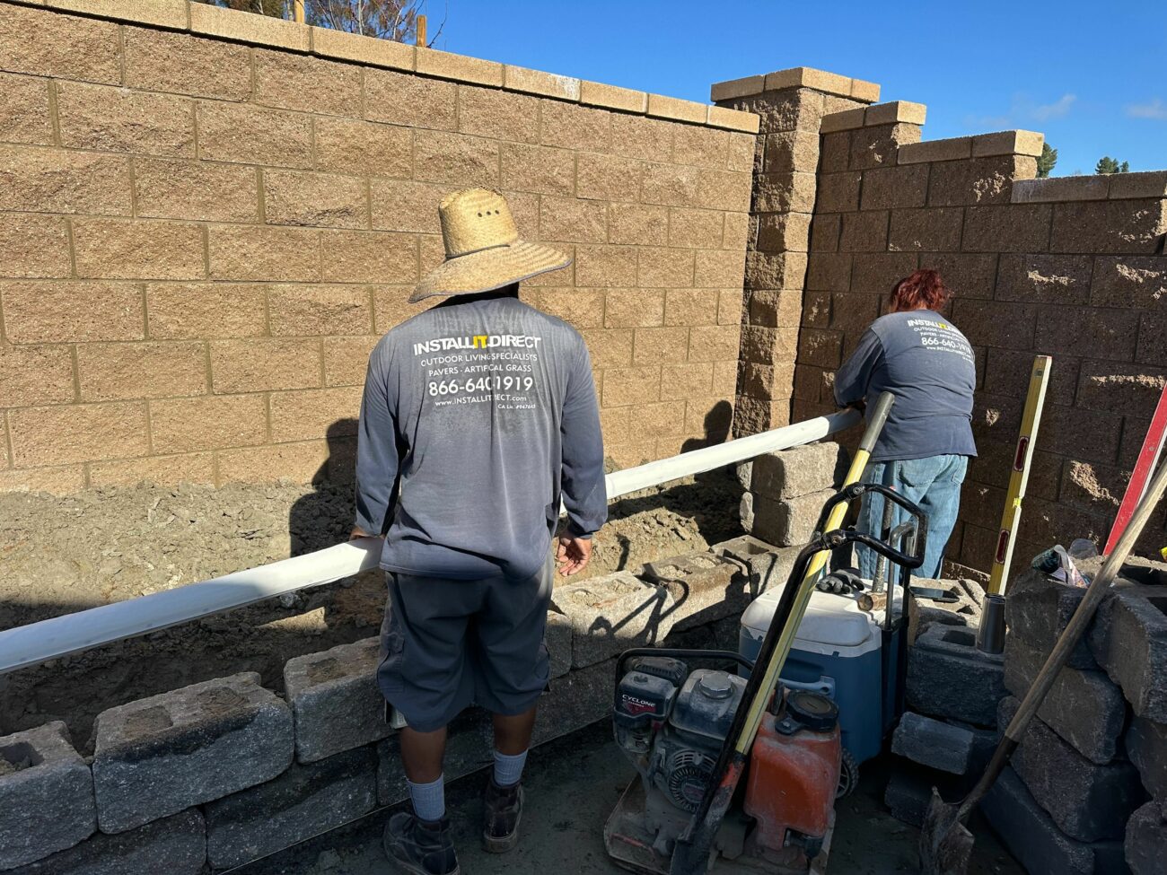 Two contractors in Install It Direct shirts lay retaining wall block, using a level and plate compactor on a sunny day.