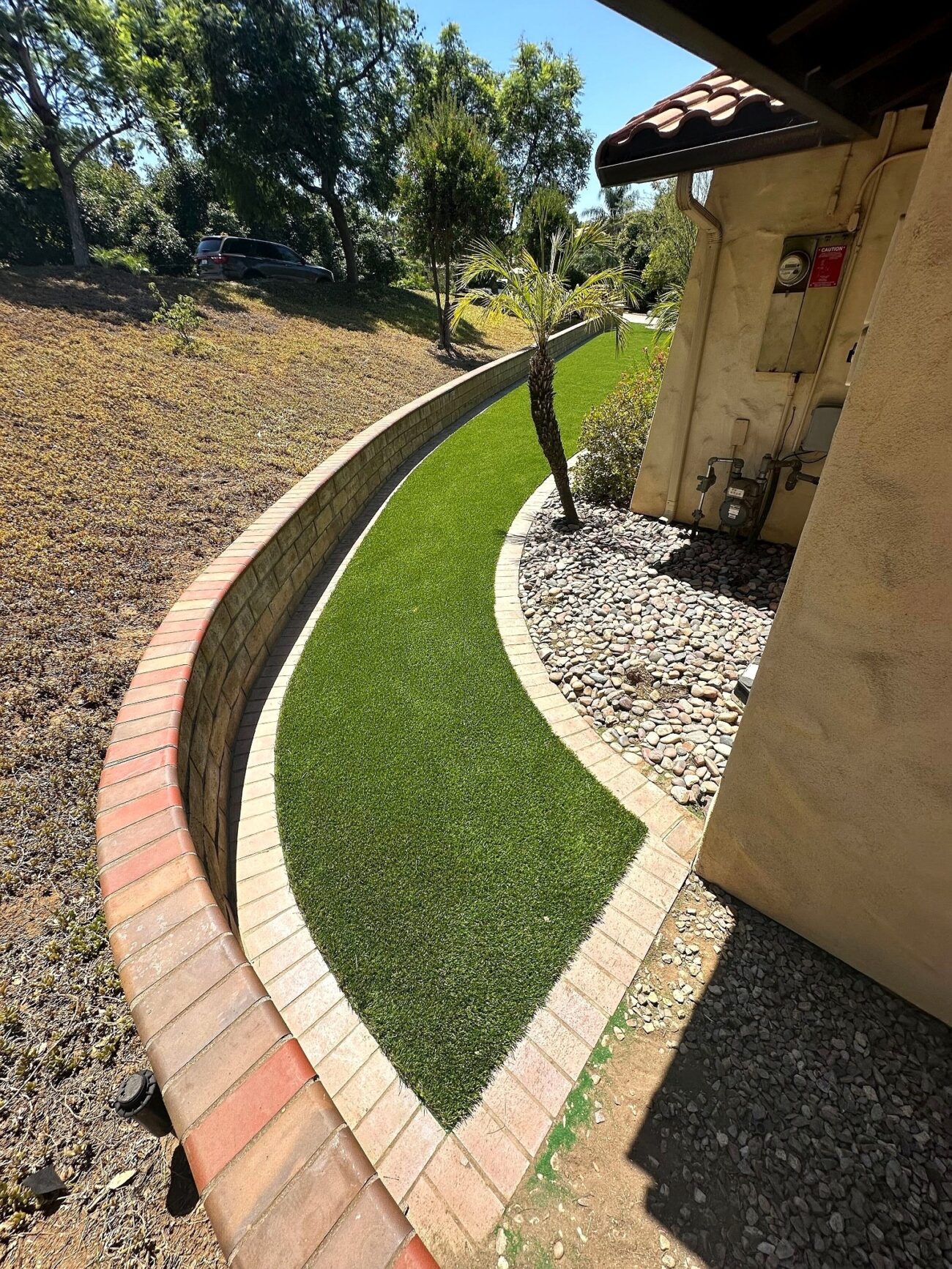 Curved synthetic turf strip bordered by brick edging winds alongside a stucco building, with river rock on one side and a...