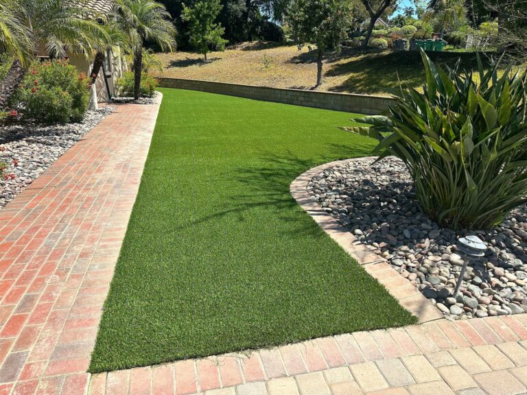 Freshly installed artificial turf lawn bordered by brick pavers and river-rock planting beds in a residential backyard.