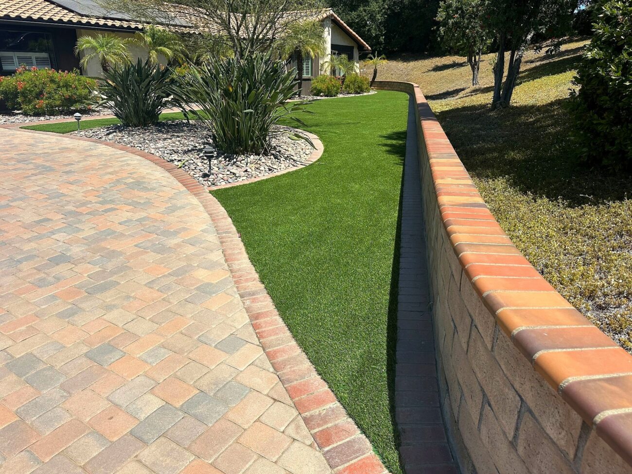 Narrow strip of synthetic turf between a curved brick paver driveway and a terracotta-capped block retaining wall, with a...