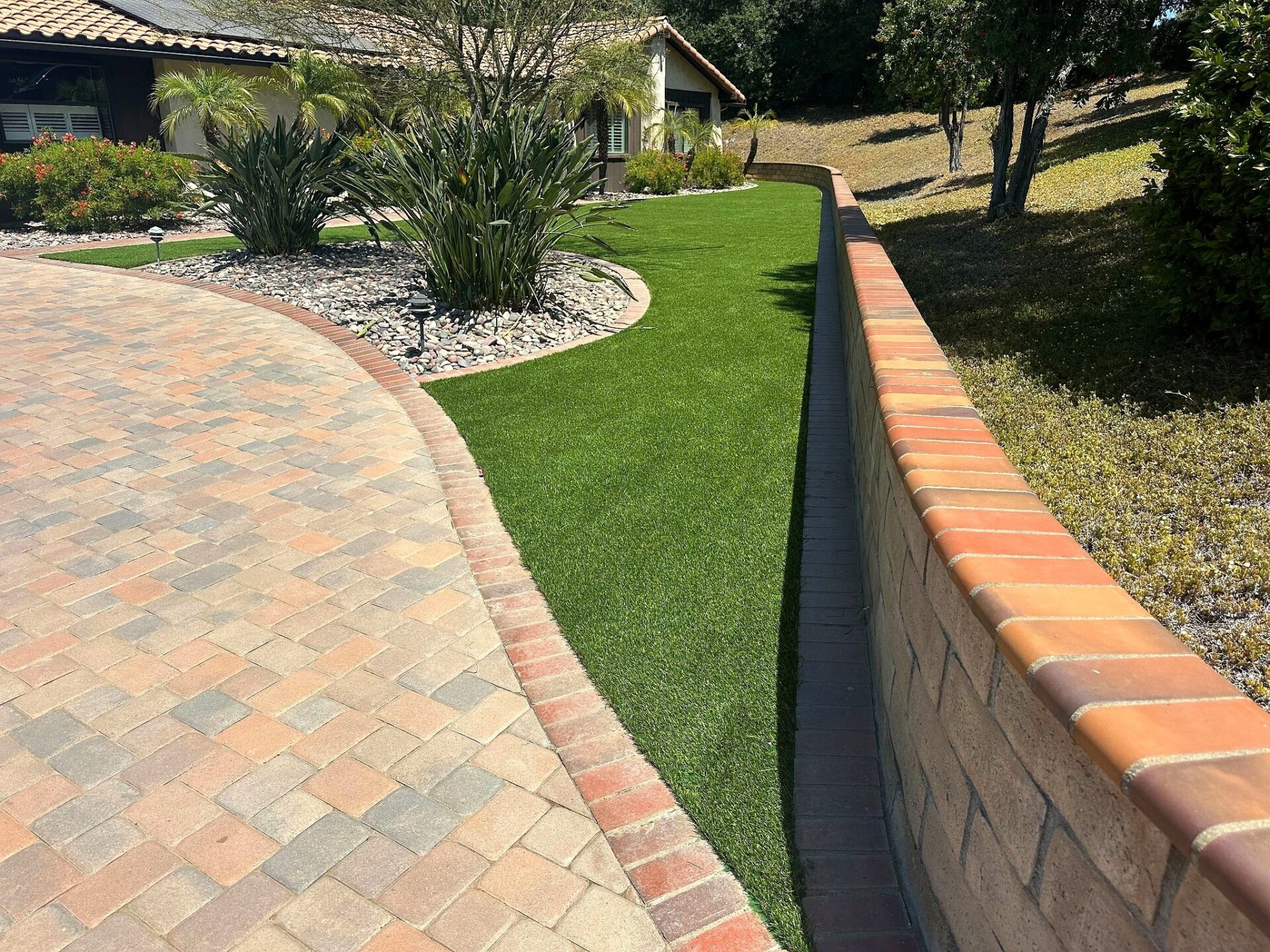 Narrow strip of synthetic turf between a curved brick paver driveway and a terracotta-capped block retaining wall, with a...