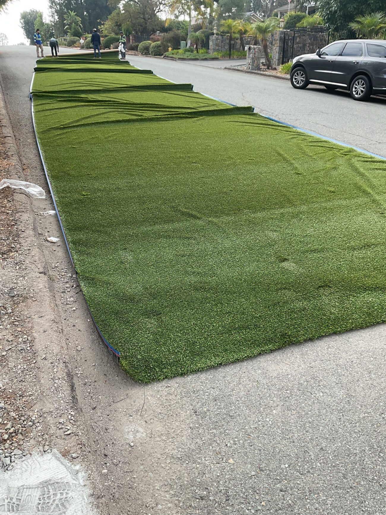 Large rolls of synthetic turf laid flat along a residential street curb, with workers visible in the background.