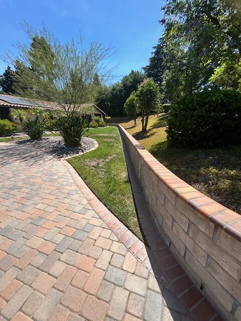 Curved brick paver driveway runs alongside a block retaining wall, with green lawn and trees beyond.