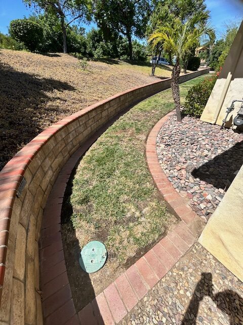 Curved brick retaining wall and paver path separating a grass lawn from a river-rock planting bed beside a stucco home.