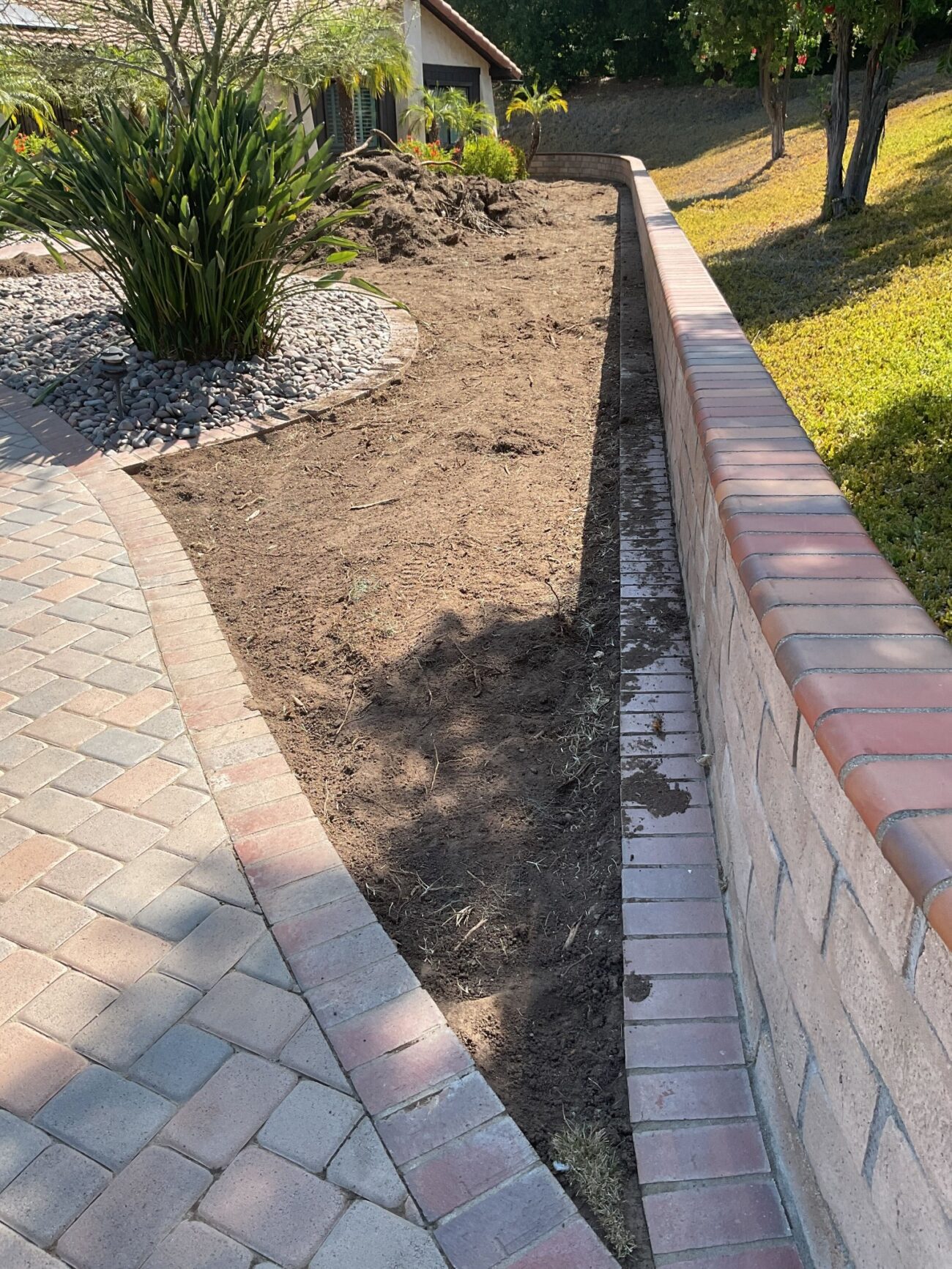 Bare soil planting bed alongside a brick retaining wall and paver driveway, cleared and ready for installation.