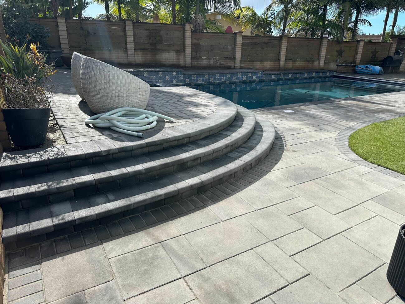 Finished backyard pool deck and patio featuring interlocking pavers with a three-step curved bullnose stair leading down to the pool. A small artificial turf lawn and a block wall/retaining wall frame the area; pavers appear clean and the installation looks complete.